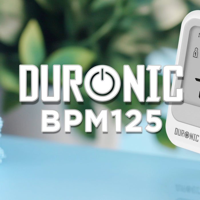 Duronic digital blood pressure monitor displaying blood pressure and pulse readings, pictured beside folded towels in a home setting.