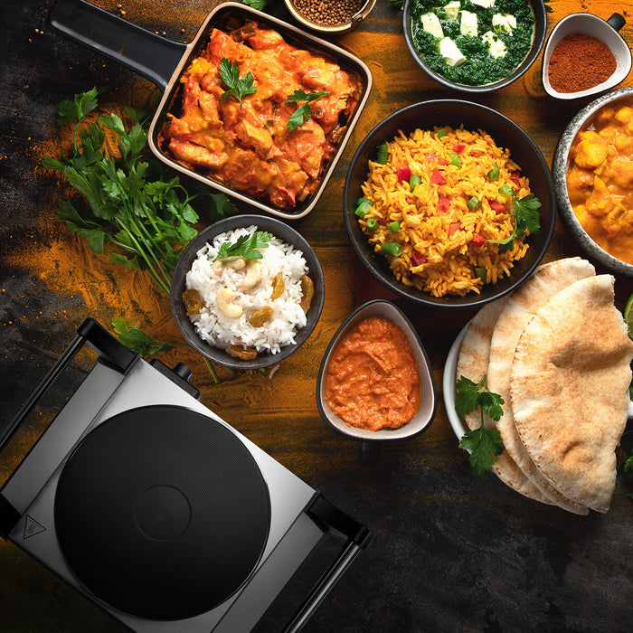 Duronic HP1 SS portable electric hot plate surrounded by indian dishes like butter chicken, rice, naan, biryani