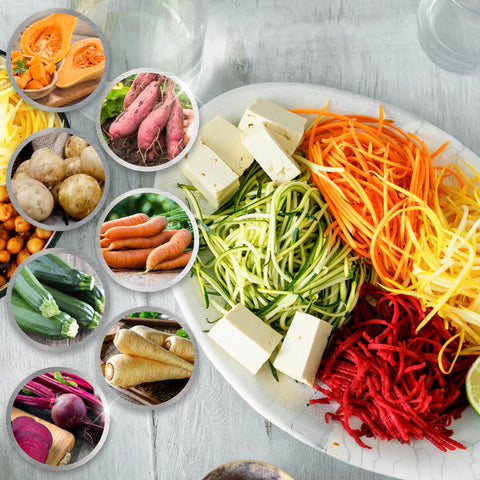 A vibrant assortment of spiralized vegetable noodles including zucchini, carrot, and beet served on a white platter with cubed tofu and circular insets showcasing the fresh whole vegetables.