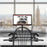 Duronic DM05D5 Corner Sit-Stand Desk with a dual-monitor mount is displayed on a white desk in a bright, modern office overlooking a city skyline.