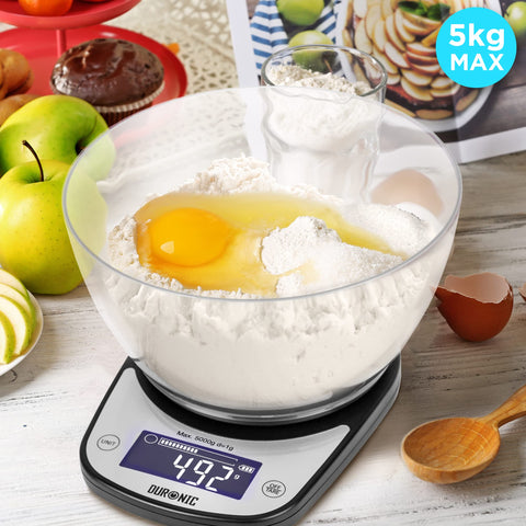 Duronic Digital Kitchen Scale weighing ingredients in a clear bowl with a 5kg maximum capacity, surrounded by baking supplies like eggs, flour, and fresh apples.