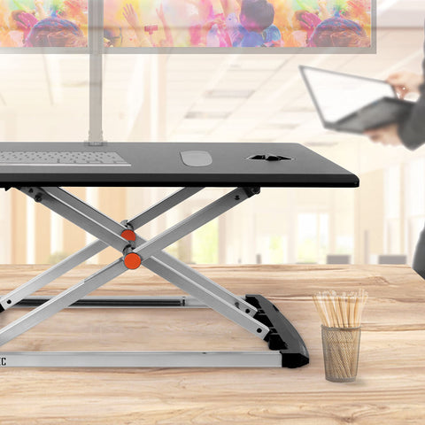 Duronic Sit-Stand Desk sleek black and silver adjustable standing desk converter sits on a wooden tabletop in a bright office setting