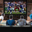 Four men watching an American football game on a large home theater projector screen.